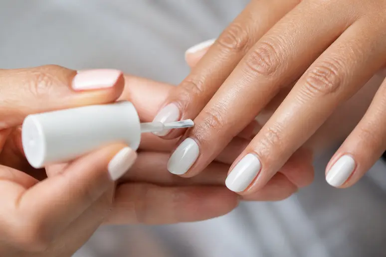 Application minutieuse d'une couche de vernis blanc opaque avant la pose du vernis ongle rose fluo.
